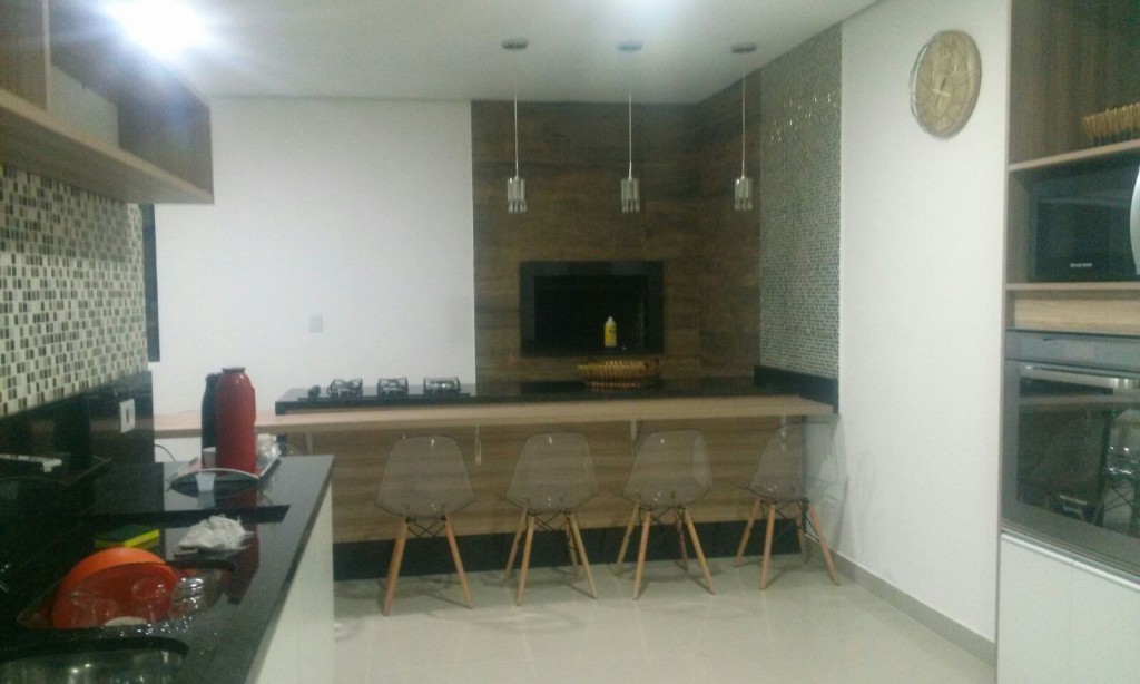 Uma moderna churrasqueira, ao fundo, tem à sua frente a ilha gourmet com fogão e bancada acoplados, além de modernas cadeiras em acrílico. Na parede à direita, o forno de microondas.